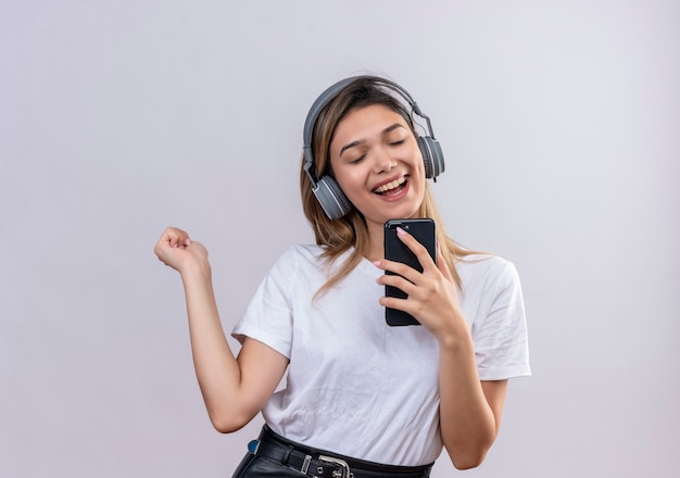 A joyful young woman in white t-shirt wearing headphones singing while listening to the music on her phone on a white wall