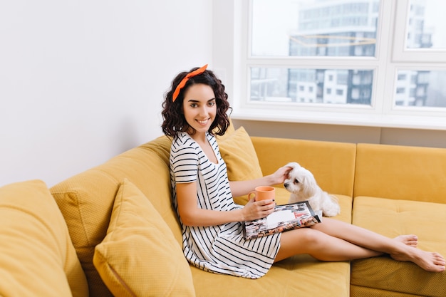 Joyful young woman with cut brunette hair in dress chilling with dog on couch in modern apartment. Reading magazine, cup of tea, comfort, cozy time at home with pets