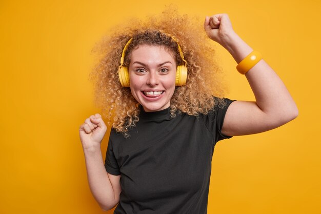 Joyful young woman with natural curly hair raises arms dances carefree enjoys favorite music in headphones has fun dressed in black t shirt isolated over yellow wall. Entertainment concept