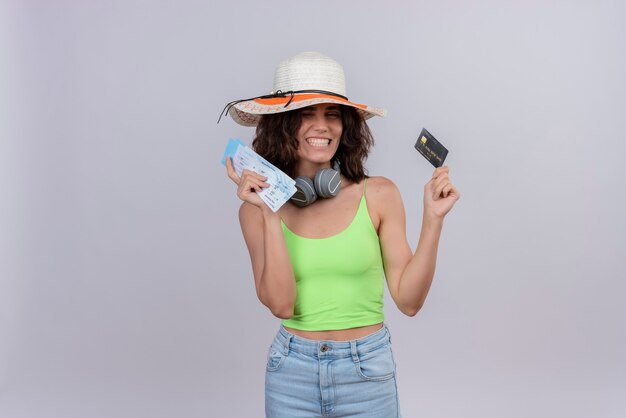 A joyful young woman with short hair in green crop top in headphones wearing sun hat smiling and holding plane tickets and credit card on a white background