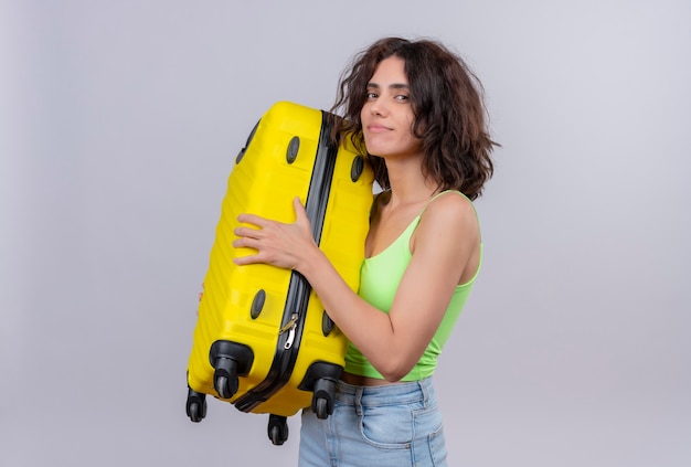 A joyful young woman with short hair in green crop top holding yellow suitcase on a white background