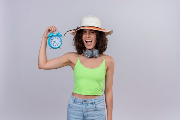 A joyful young woman with short hair in green crop top wearing sun hat holding blue alarm clock on a white background