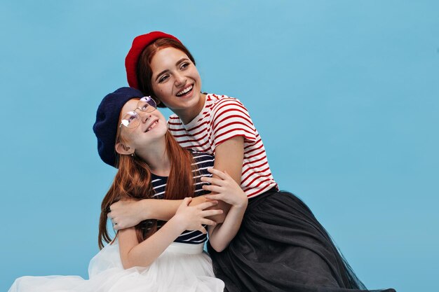 Joyful young women with lovely smile in bright cool berets and black and white bouffant skirts hugging and sitting on floor