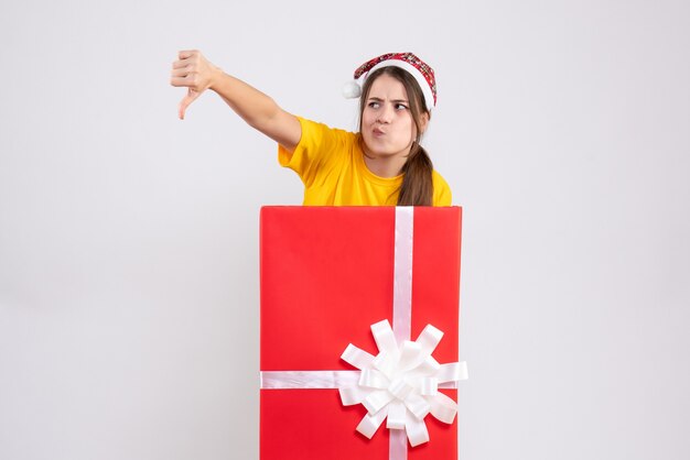joyless girl with santa hat making thumb down sign standing behind big xmas gift on white