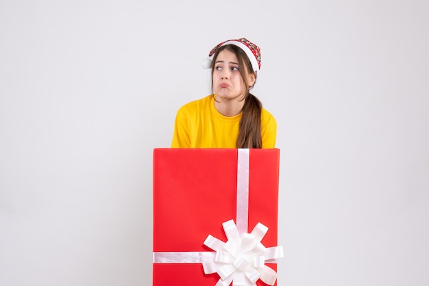 joyless girl with santa hat standing behind big xmas gift on white