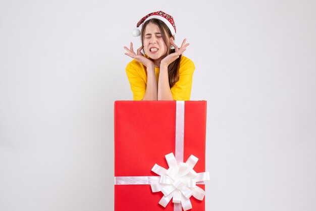 Free photo joyless girl with santa hat standing behind big xmas gift on white