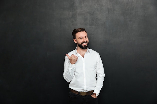 Joyous adult guy in office posing on camera, gesturing with thumb aside over dark gray copy space