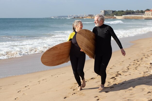 Joyous aged couple on beach with surfboards. Smiling gray-haired man and woman in wetsuits spending summer vacation near sea training and surfing. Active life, health care of mature people concept