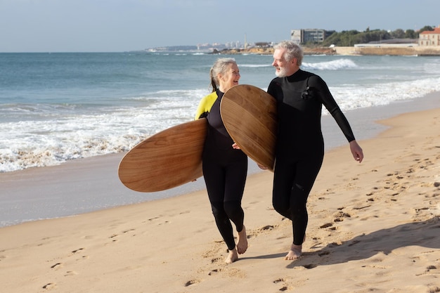 サーフボードとビーチで楽しい老夫婦。海のトレーニングやサーフィンの近くで夏休みを過ごすウェットスーツで笑顔の白髪の男性と女性。アクティブな生活、成熟した人々の健康の概念の概念