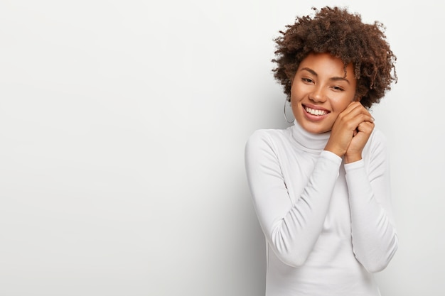 Joyous black woman with pleasant smile, keeps hands together near face, looks happily