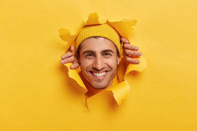 Joyous Caucasian man with toothy smile, shows white teeth, looks through hole torn in paper