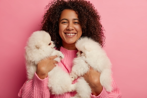 Foto gratuita gioiosa ragazza dalla pelle scura riposa con due cani a casa, porta due soffici cuccioli di razza spitz