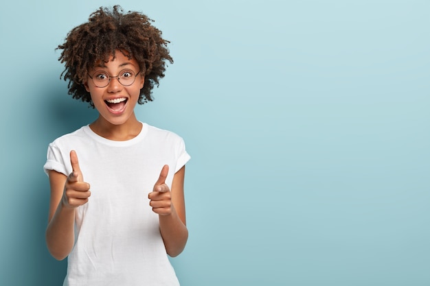 Joyous dark skinned woman points both fore fingers straightly at camera