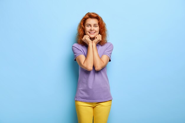 Joyous ginger woman holds chin, smiles gently, being in good mood