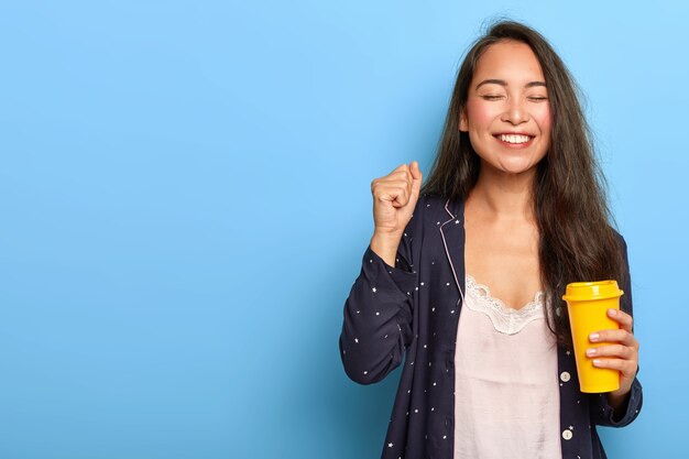 Joyous happy Asian woman with long dark hair, enjoys morning coffee, clenches fist, dressed in nightwear, smiles broadly