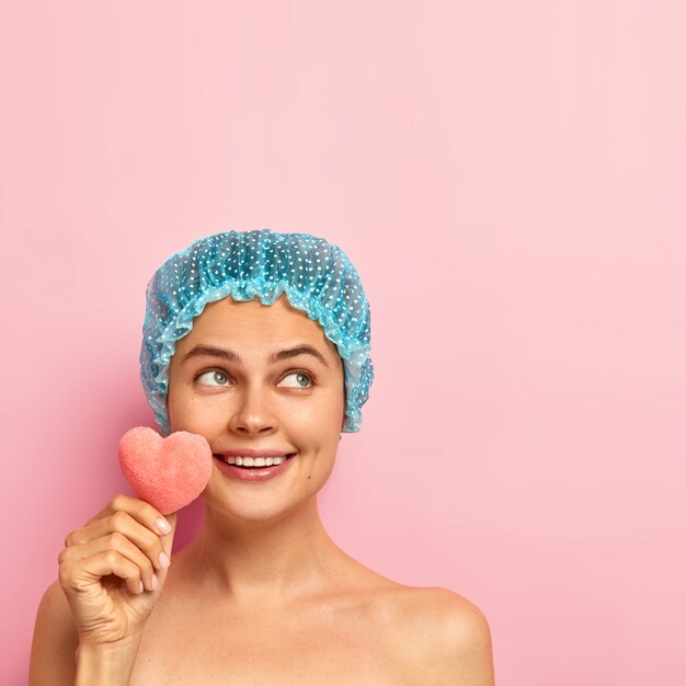 Joyous happy young woman has well cared skin, toothy smile, stands with dreamy expression, holds small cosmetic sponge, wears protective showercap