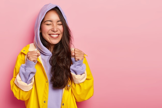 Joyous lovely woman raises clenched fists, rejoices awesome stroll with boyfriend during autumn day, dressed in violet sweatshirt with hoody and yellow raincoat