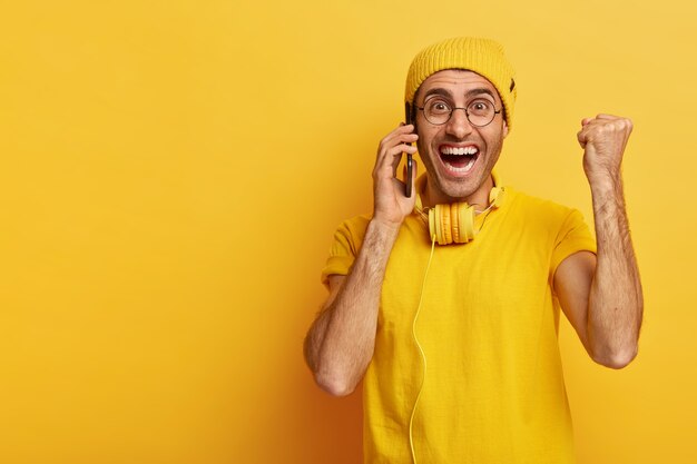 Joyous optimistic man with toothy smile, raises clenched fist in triumph, calls via smartphone, enjoys conversation