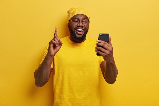 즐거운 통통한 남자가 검지 손가락을 들고 휴대 전화를 들고 인터넷 서핑을위한 여가 시간을 즐기고 노란색 모자와 캐주얼 티셔츠를 입습니다.