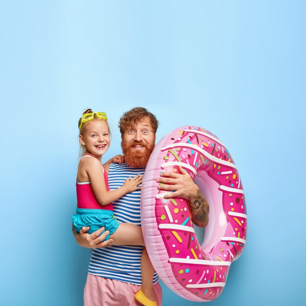 Joyous red haired family have fun at coastline. Pleased cheerful bearded dad holds little girl and inflated swim ring