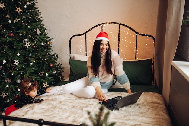 Joyous woman sitting on bed while looking at laptop screen while child standing near Christmas tree