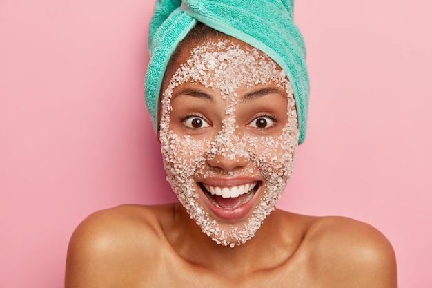 Joyous woman smiles positively has brows eyes, well cares body skin, wears sea salt scrub for cleansing complexion