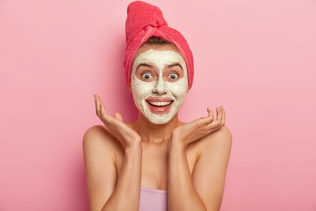 Joyous young lady with cheerful expression, spreads palms near face, applies natural clay mask for looking refreshed, has charming smile, wrapped in soft bath towel