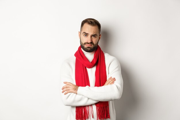 Judgemental adult man in christmas scarf and sweater looking skeptical at camera, dislike something, standing over white background.