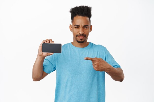 Judgemental african american man furrow eyebrows, pointing finger at smartphone screen with disapproval, dislike app, bad interface application, standing against white background