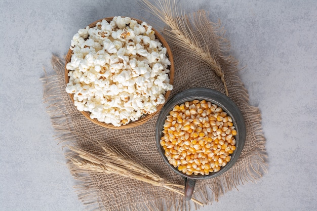 Jug filled with corn and bowl filled with popcorn next tot stalks of wheat on a piece of cloth on marble.