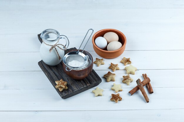 Jug of milk, bowl of flour, flour strainer in a wooden board with star cookies, cinnamon, eggs close-up on a white wooden board background