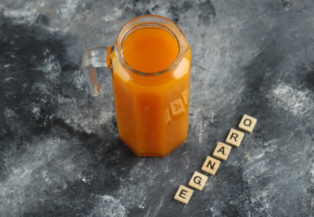 Jug of orange juice with wooden letters on marble table.