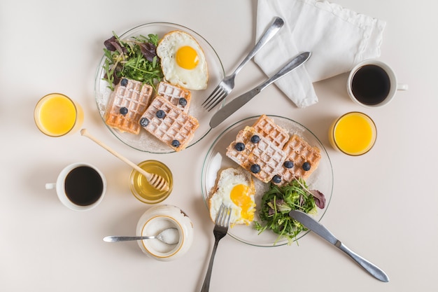 Juice glass; tea cup; honey; powdered milk and plate of waffles; fried eggs with salad on white backdrop