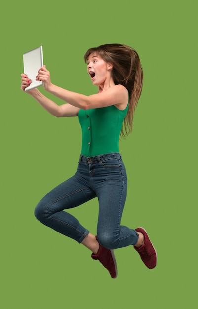 점프하는 동안 노트북 또는 태블릿 가제트를 사용하여 녹색 스튜디오 배경 위에 젊은 여자의 점프. 모션 또는 움직임에서 Runnin 소녀. 인간의 감정과 표정 개념. 현대 생활의 가제트