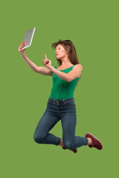 Прыжок молодой женщины над зеленой предпосылкой студии используя гаджет компьтер-книжки или планшета во время прыжка. Бегущая девушка в движении или движении.