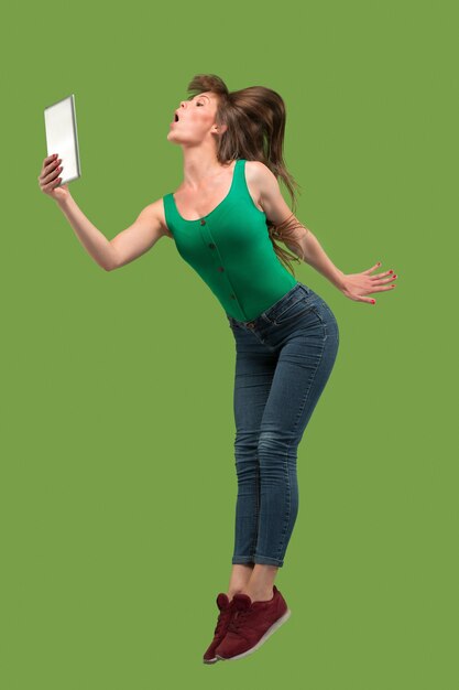 Jump of young woman over green studio background using laptop or tablet gadget while jumping.