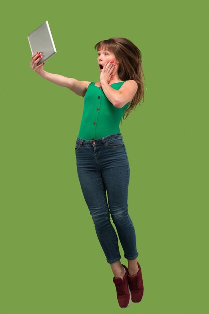 Jump of young woman over green studio background using laptop or tablet gadget while jumping.