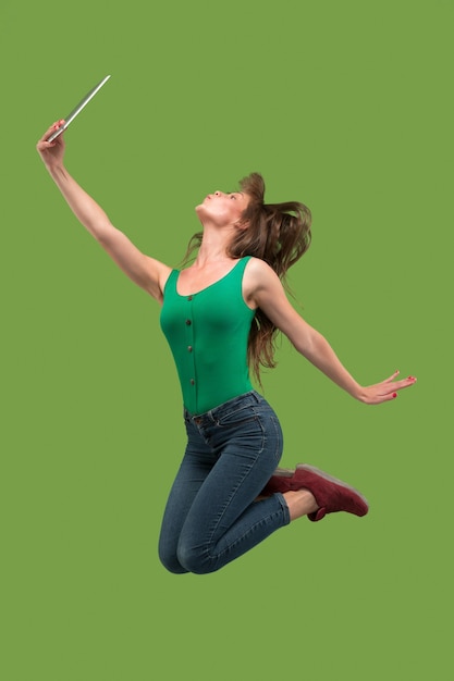 Jump of young woman over green studio background using laptop or tablet gadget while jumping