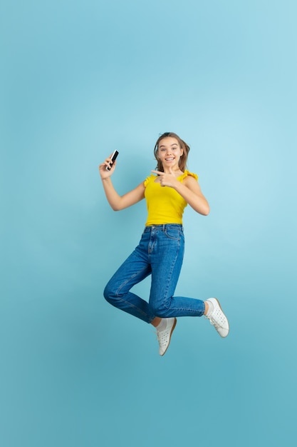 Jumping high with smartphone. Caucasian teen girl's portrait on blue