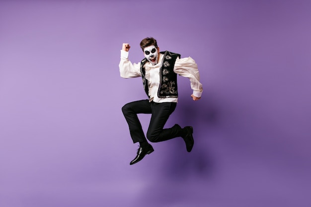 ハロウィーンの衣装で笑う男をジャンプします。紫色の壁に踊るメキシコの化粧をした興奮した男の屋内写真。