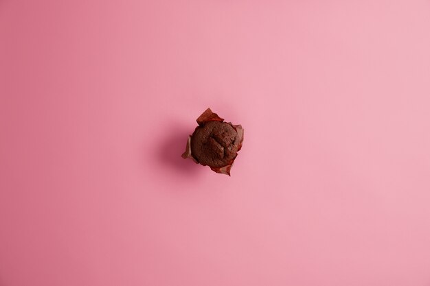 上から撮影した茶色の紙においしい甘いチョコレートマフィンを焼き、ピンクの背景に分離して、すぐに食べられます。ジャンクフード、デザート、カロリー、栄養の概念。セレクティブフォーカス