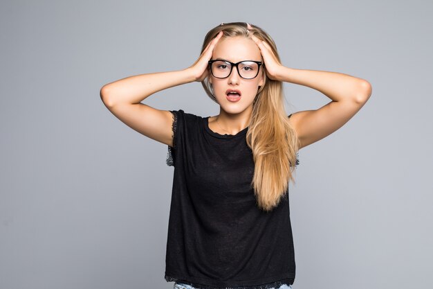 Just happy. Young smiling exited woman in casual wear holding hands on head and shouting while standing against gray studio background. Cheerful crazy girl with open mouth posing to camera