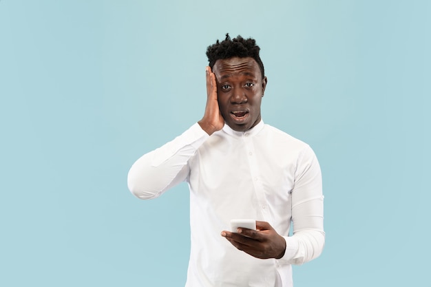 Just lost a bet. Young african-american man with smartphone isolated on blue studio background, facial expression. Beautiful male half-lenght portrait. Concept of human emotions, facial expression.