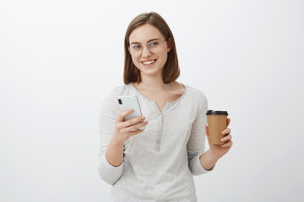Just sec need answer. Charming carefree nice european woman in glasses holding paper cup with coffee drinking beverage enjoying talk with boss and typing message in smartphone over gray wall