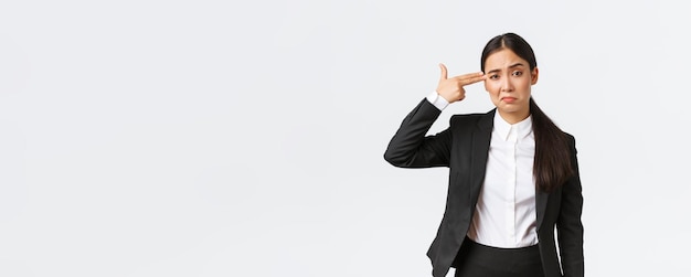 Just shoot me please Bothered and fed up asian saleswoman hate her work standing in business suit shooting herself with hand gun gesture as feeling exhausted standing white background