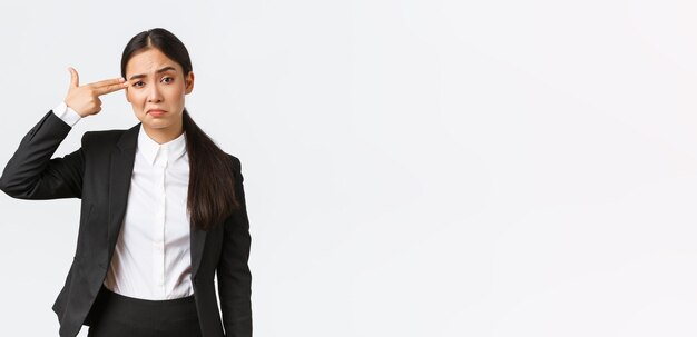 Just shoot me please Bothered and fed up asian saleswoman hate her work standing in business suit shooting herself with hand gun gesture as feeling exhausted standing white background