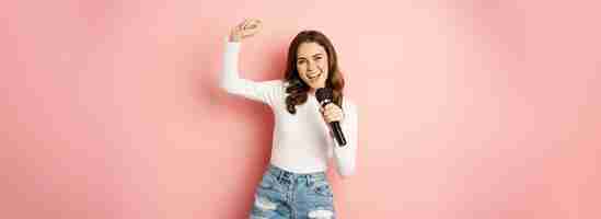 Бесплатное фото Караоке счастливая улыбающаяся девушка поет в микрофоне, танцует и веселится, стоя над розовой спинкой