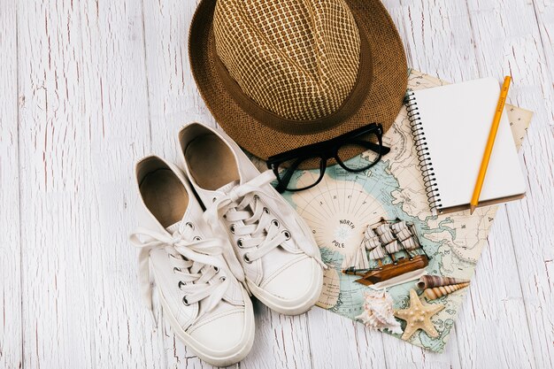 Keds, hat, glasses, notebook, little wooden ship lie on white travel map