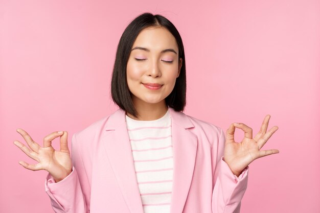 침착하게 웃고 있는 젊은 아시아 여성이 직장에서 요가 마음챙김과 휴식을 명상하며 분홍색 배경 위에 서서 기뻐하는 얼굴로 공기를 들이마십니다.