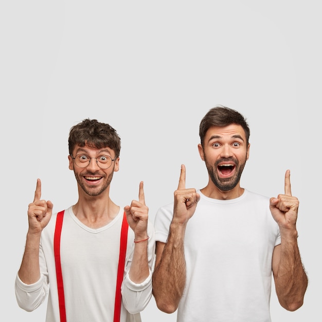 Keep heads up! Optimistic unshaven brothers point with both index fingers, smile broadly while show new banner, dressed in white clothes, isolated over  wall, demonstrate new amazing product